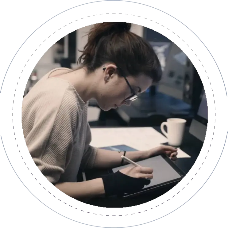 A person with dark hair tied up in a bun, wearing glasses and a light sweater, works on a digital tablet with a stylus in a dimly lit room. A mug and monitors are in the background. They appear focused and are wearing a wrist brace on one hand.