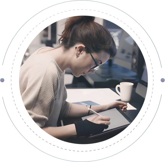 A person with dark hair tied up, wearing glasses and a beige sweater, works on a digital tablet with a stylus pen. They are seated at a desk with a computer monitor and a white mug in the background, appearing focused on their task.